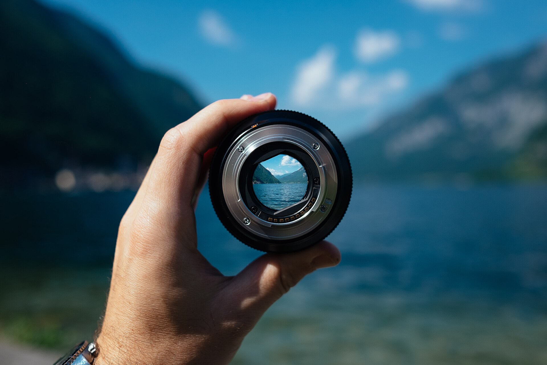 A hand holding a camera lens that frames a mountain lake